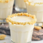 Close up side view of Salted Nut Roll shot garnished with crushed peanuts and salt on the rim. More Salted Nut Roll shots on counter, along with peanuts and salt.