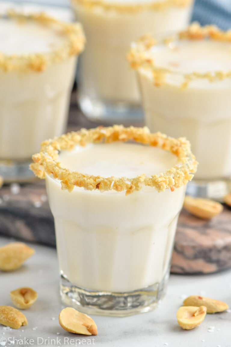 Close up side view of Salted Nut Roll shot garnished with crushed peanuts and salt on the rim. More Salted Nut Roll shots on counter, along with peanuts and salt.