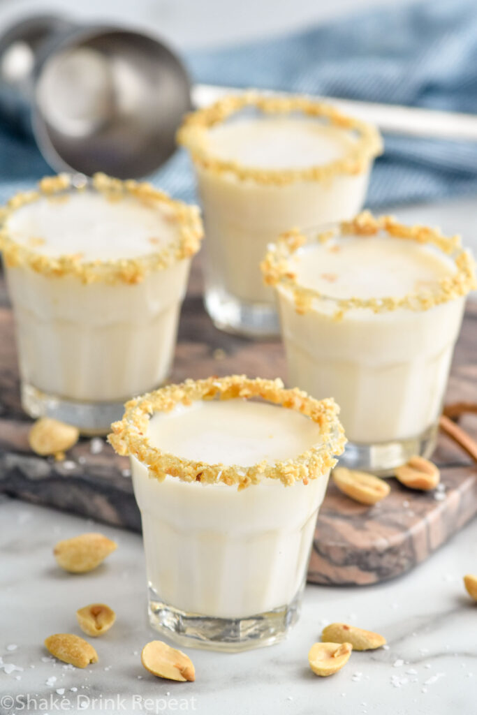 Side view of four Salted Nut Roll shots garnished with crushed peanuts on the rim. Cocktail jigger in the background. Extra salt and peanuts beside shot glasses.