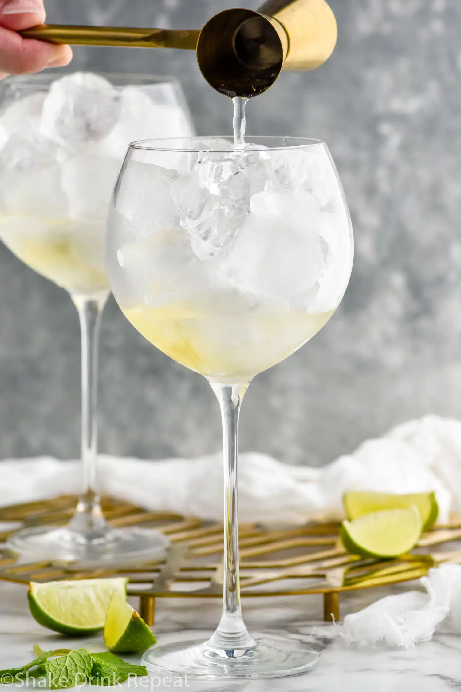 hugo spritz ingredients pouring into a wine glass with ice. Mint leaves and lime slices sitting beside glass.