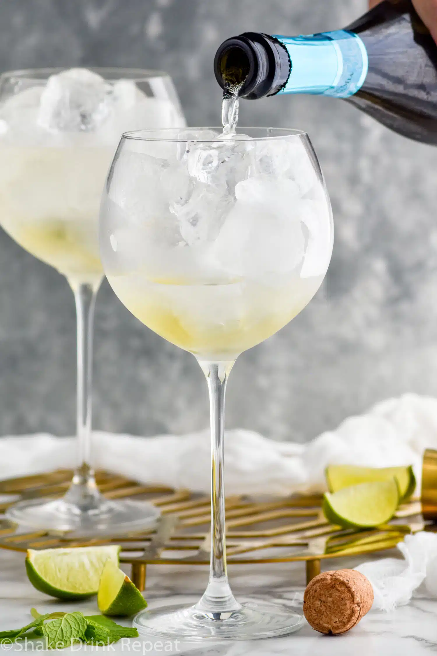 bottle of sparkling wine pouring into a wine glass with ice. Mint leaves and lime slices sitting beside glass.