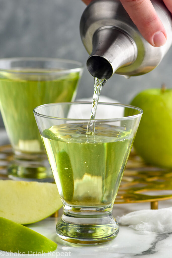 Man's hand pouring cocktail shaker of appletini ingredients into a glass. Green apple and slices sitting beside.