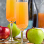 Photo of two Apple Cider Mimosas with apples, cinnamon sticks, cork, bottle of champagne, and jug of apple cider beside.