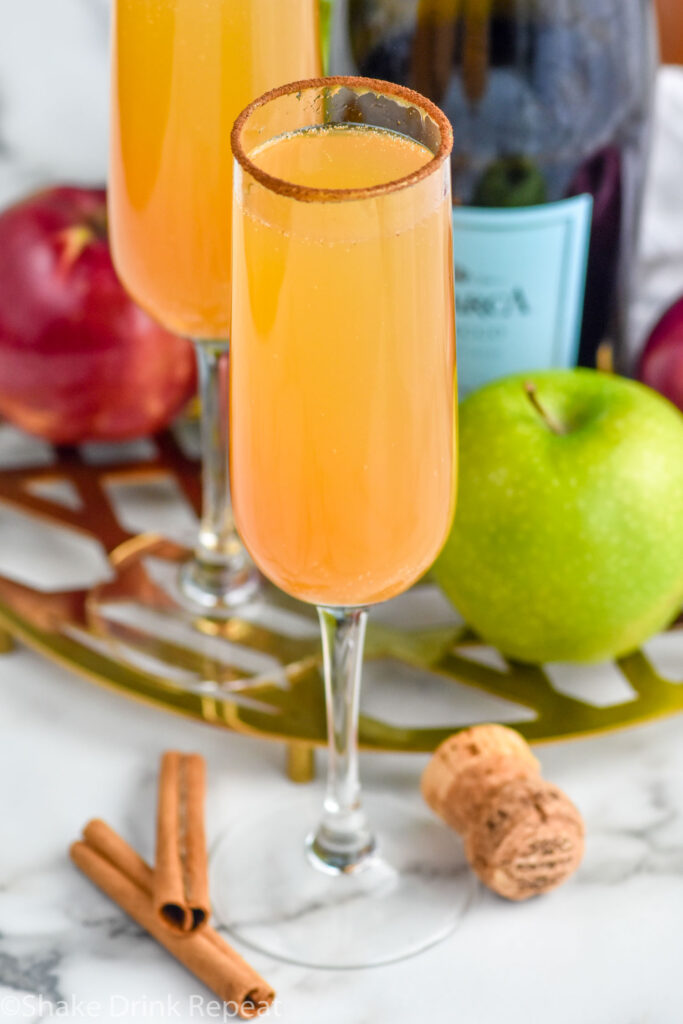 Photo of Apple Cider Mimosa with cinnamon sticks, cork, apples, and bottle of champagne beside.