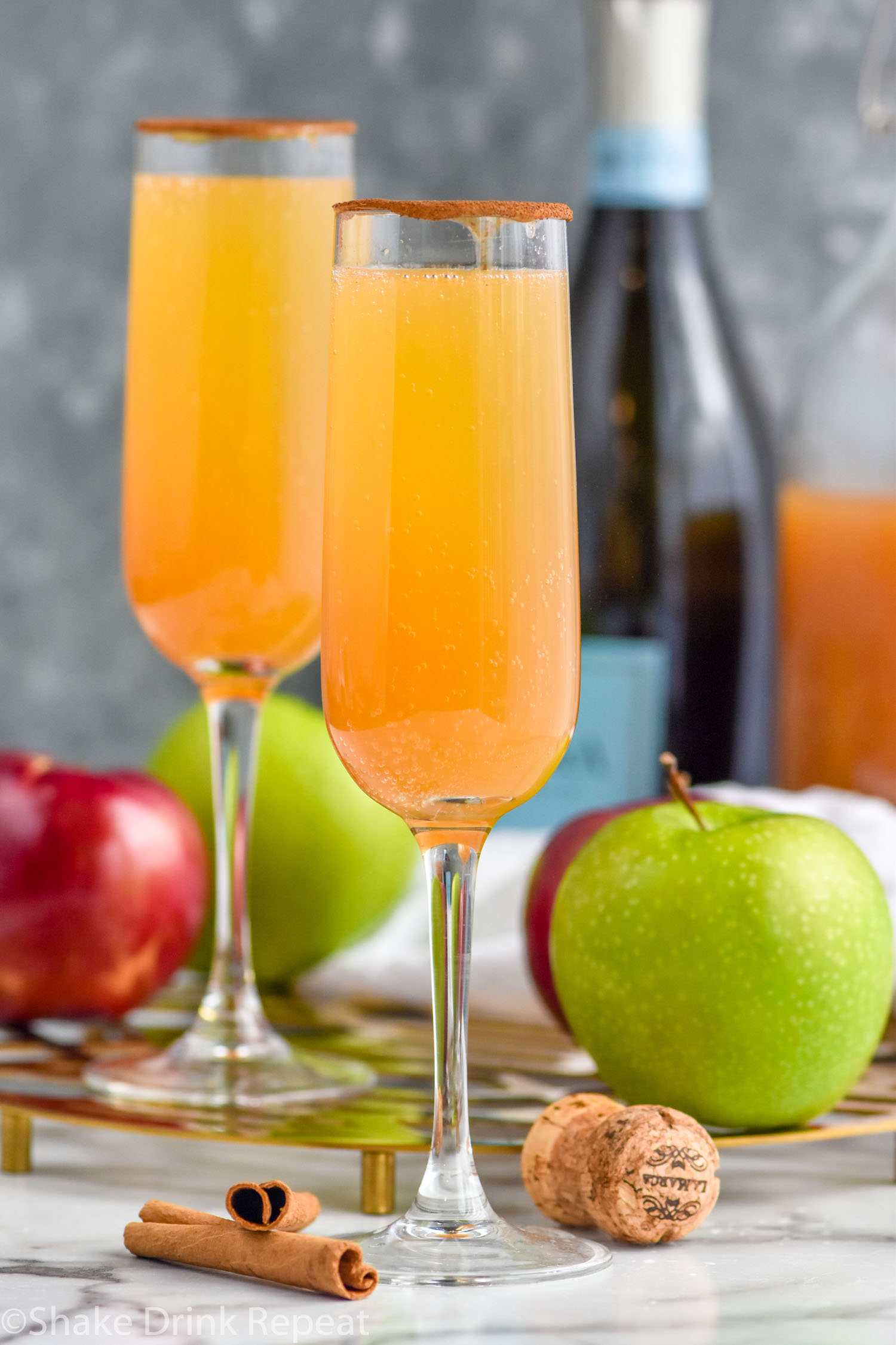 Photo of two Apple Cider Mimosas with apples, cinnamon sticks, cork, bottle of champagne, and jug of apple cider beside.