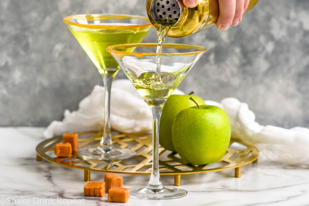 Side view of person's hand pouring cocktail shaker of Caramel Apple Martini recipe into prepared martini glasses for Caramel Apple Martini recipe. Apples and caramels on tray beside.