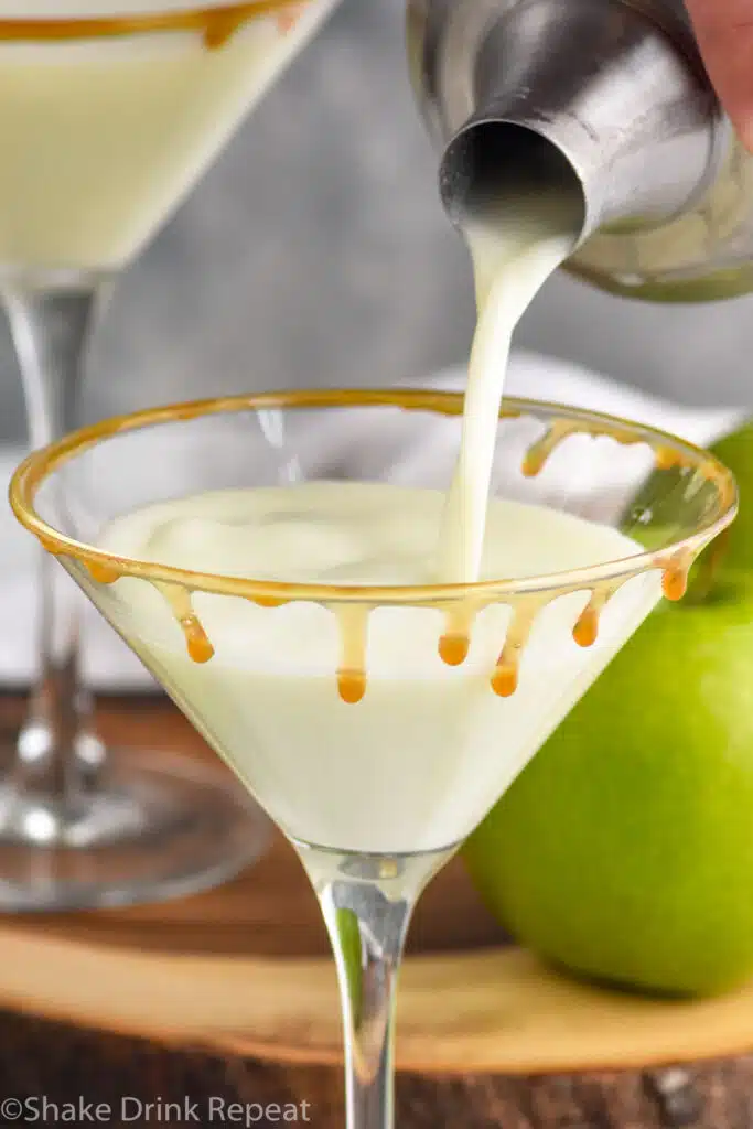 Person's hand pouring cocktail shaker of Creamy Caramel Apple Martini recipe into martini glass with caramel. Apples beside