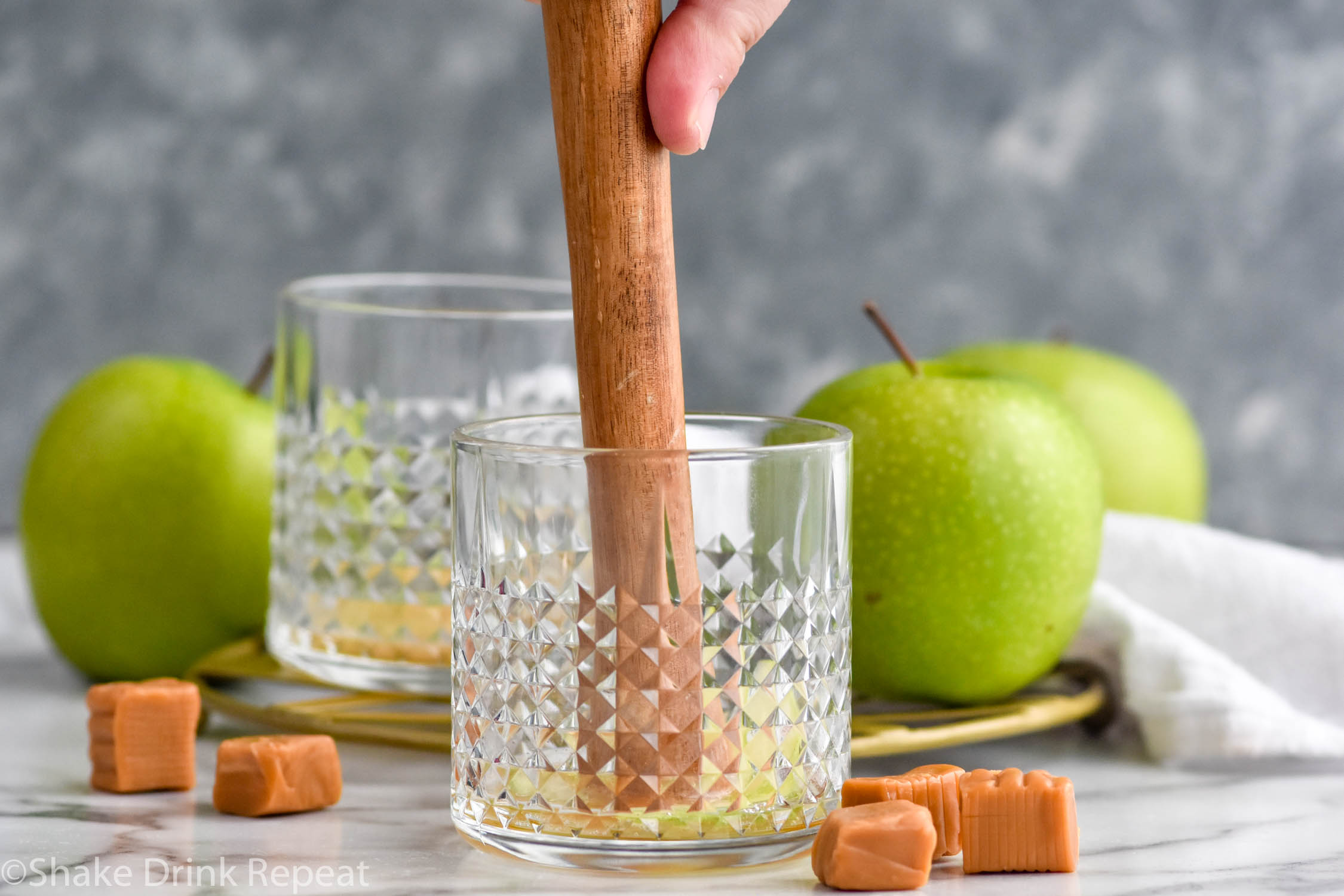 Side view of person's hand muddling ingredients for Caramel Apple Old Fashioned recipe. Apples and caramels beside glasses.
