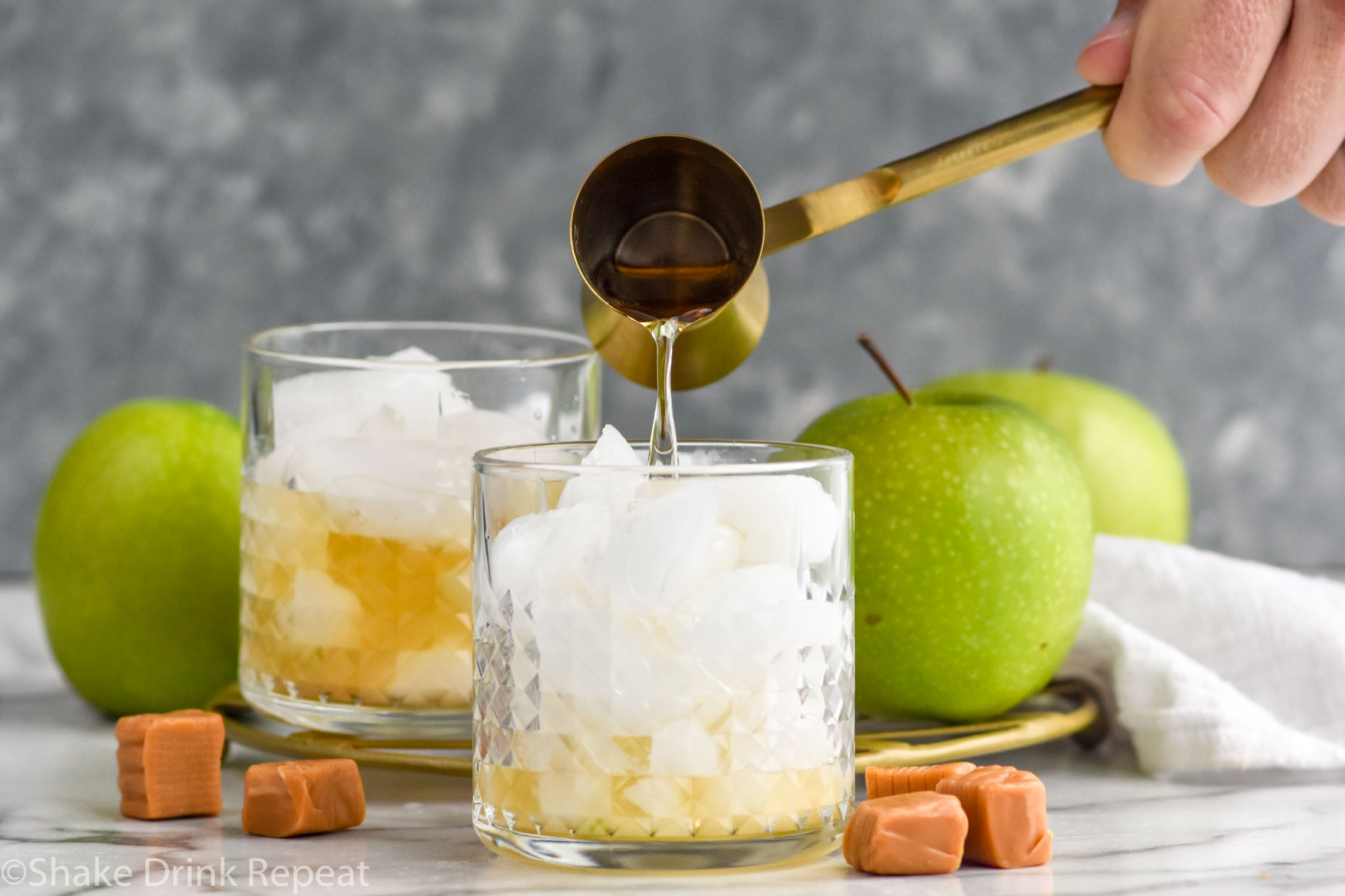 Side view of person's hand pouring cocktail jigger of ingredient into tumbler with ice and ingredients for Caramel Apple Old Fashioned recipe. Caramels and apples beside.