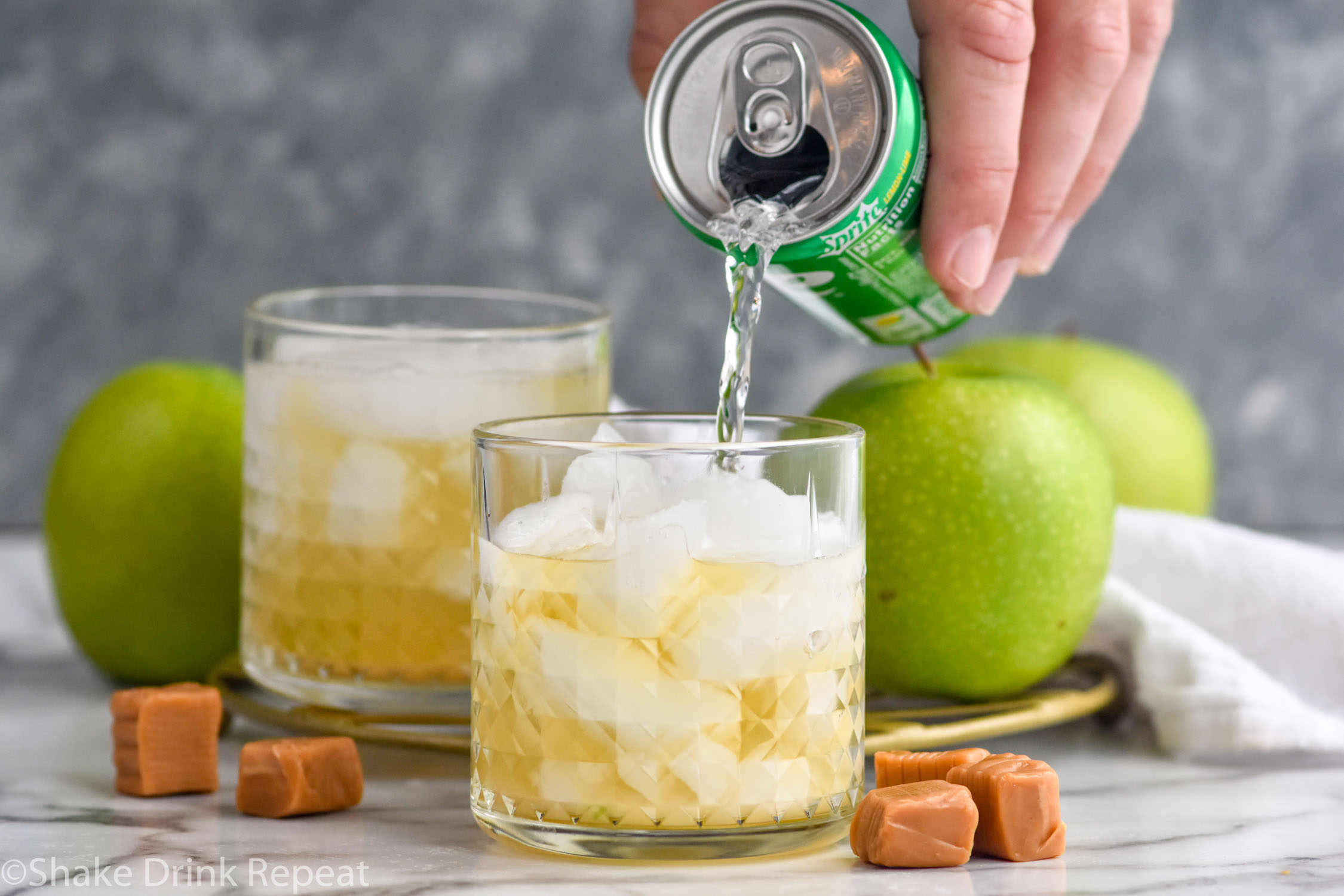 Side view of person's hand pouring lemon lime soda into glass of ingredients for Caramel Apple Old Fashioned recipe. Apples and caramels beside.