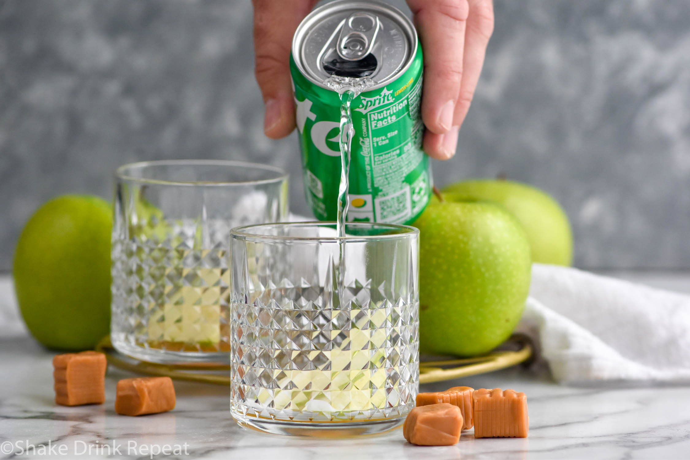 Side view of person's hand pouring lemon lime soda into glass of ingredients for Caramel Apple Old Fashioned recipe. Apples and caramels beside.