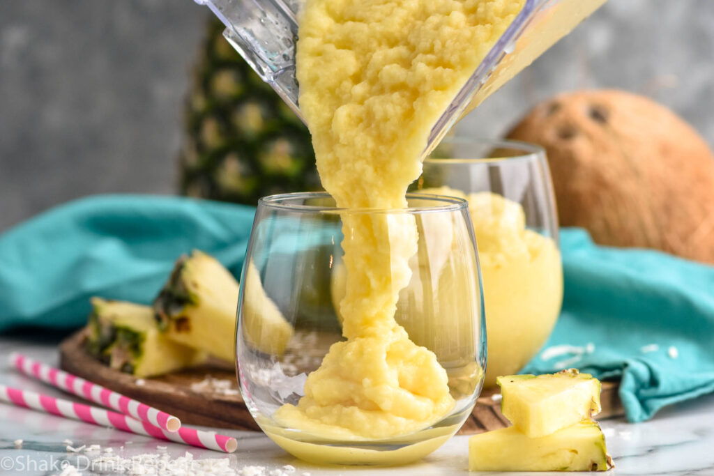 Side view of blender of Non Alcoholic Pina Colada recipe being poured into a glass. Pineapple slices beside.