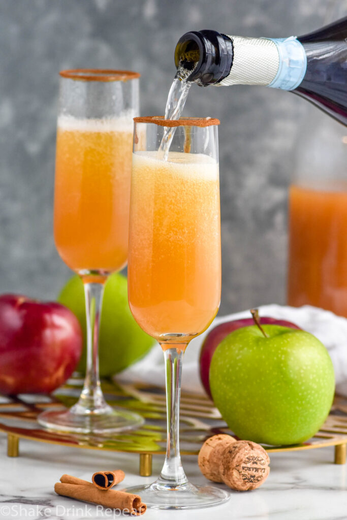 Bottle of champagne being poured into champagne glass for Apple Cider Mimosa recipe. Apples, cinnamon sticks, and cork beside.