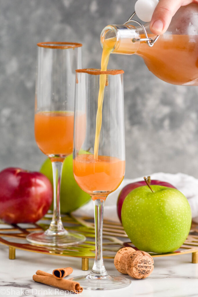 Side view of a jug of apple cider being poured into champagne glasses for Apple Cider Mimosa recipe. Apples, cinnamon sticks, and cork beside.