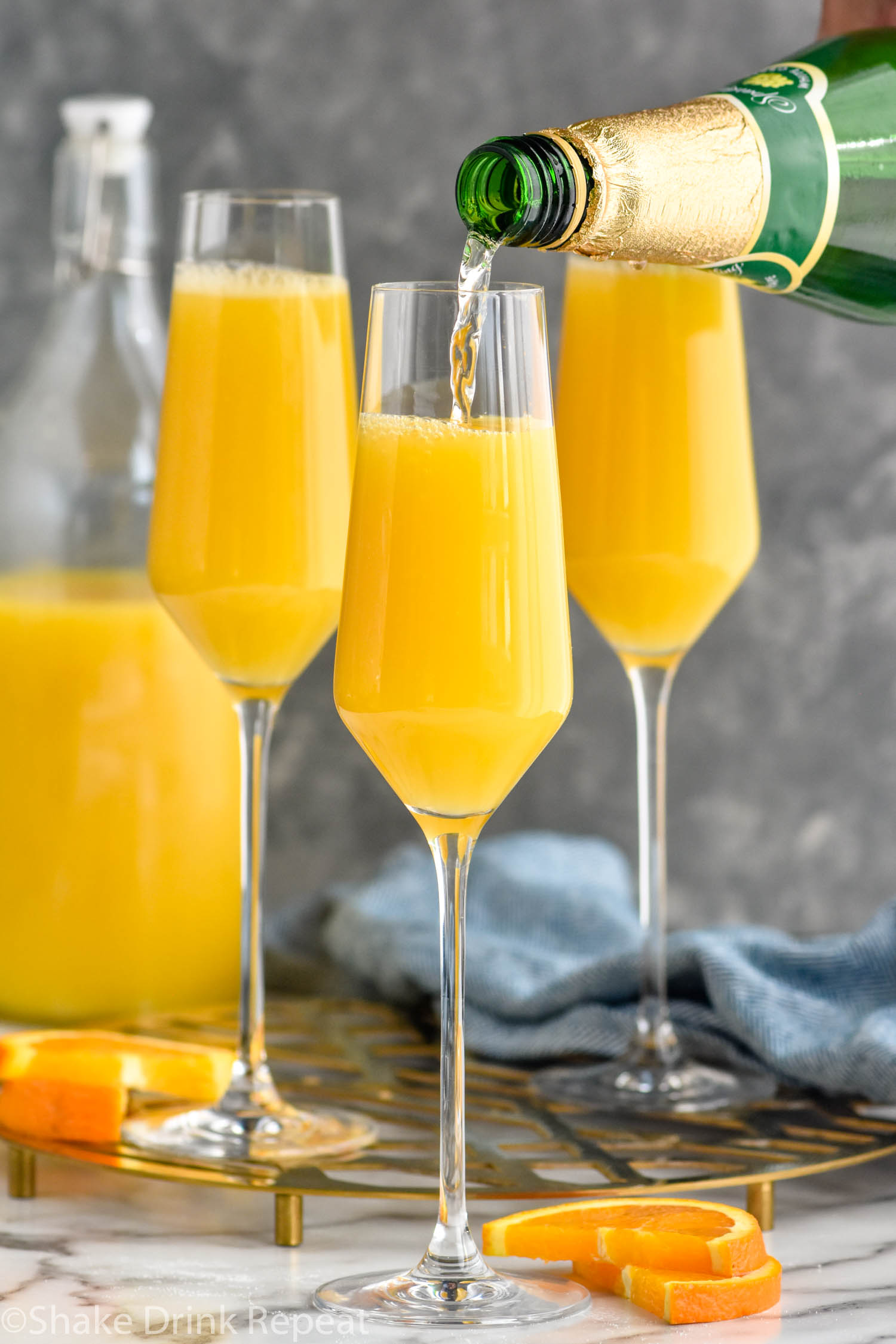 Side view of sparkling grape juice being poured into champagne glasses of orange juice for Non Alcoholic Mimosa recipe. Orange slices and bottle of orange juice beside.