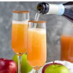 Pinterest graphic for Apple Cider Mimosa recipe. Text says, "the best Apple Cider Mimosa shakedrinkrepeat.com." Image shows bottle of champagne being poured into garnished glass for Apple Cider Mimosa recipe. Apples, cinnamon sticks, and cork beside.