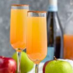Pinterest graphic for Apple Cider Mimosa recipe. Text says, "the best apple cider mimosa only uses 2 simple ingredients! shakedrinkrepeat.com" Image shows two Apple Cider Mimosas with apples and bottle of champagne beside