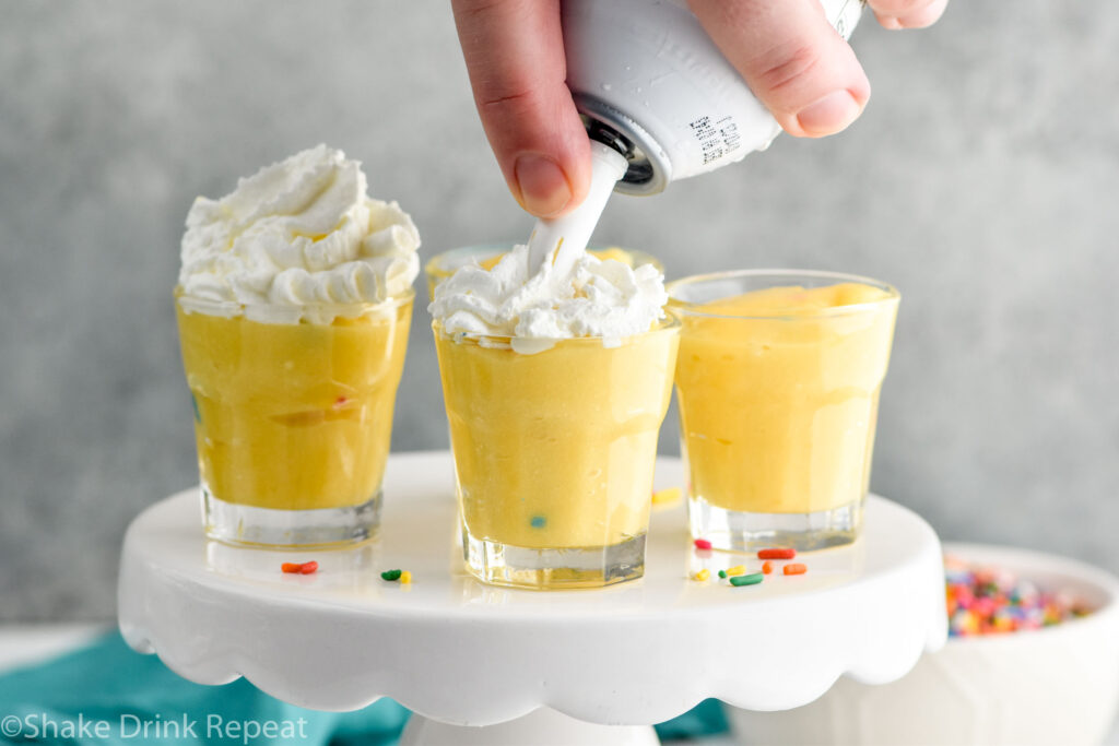 man's hand putting whipped cream on top of Birthday Cake Pudding Shots that are sitting on cake stand, surrounded by sprinkles