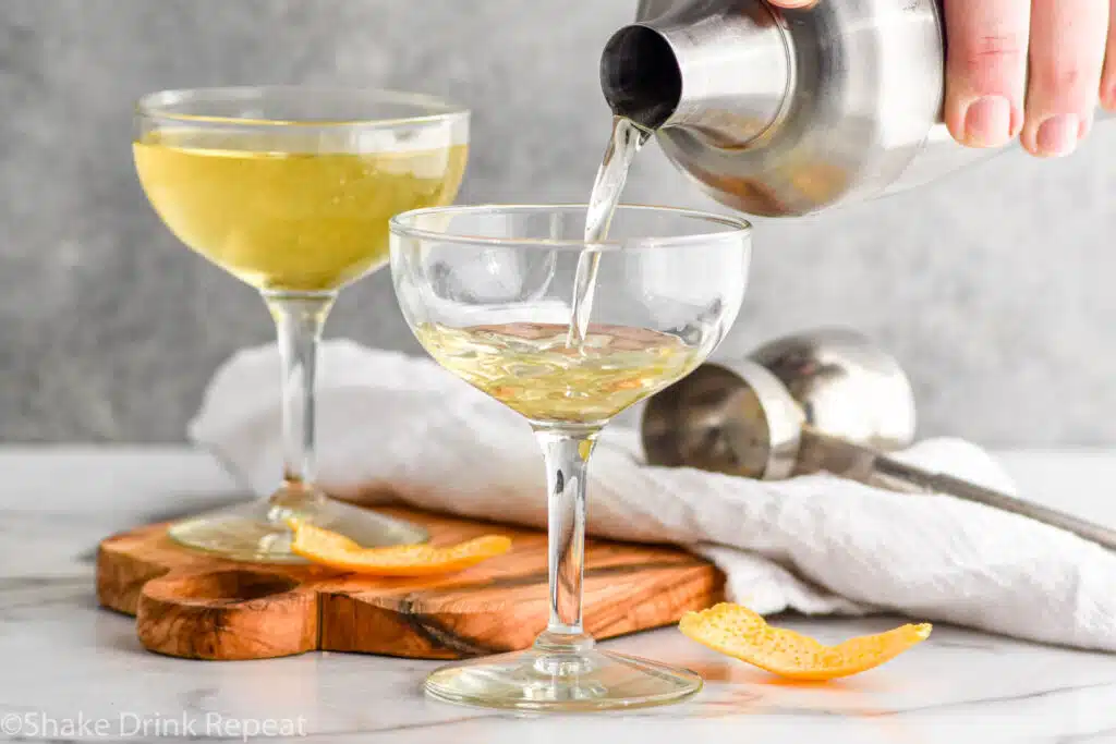 Side view of cocktail shaker of ingredients being poured into coupe glass for angel face cocktail. Orange peel beside for garnish. Another coupe glass of angel face cocktail and cocktail jigger beside.