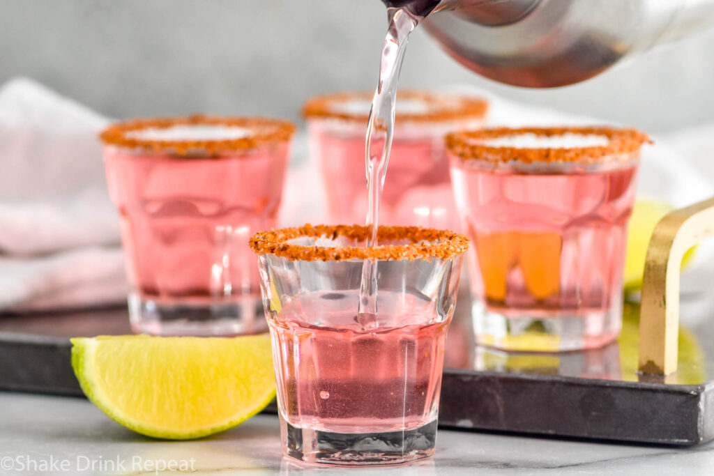 Side view of cocktail shaker of ingredients for Mexican Candy Shot recipe being poured into prepared shot glasses. Lime wedges beside.