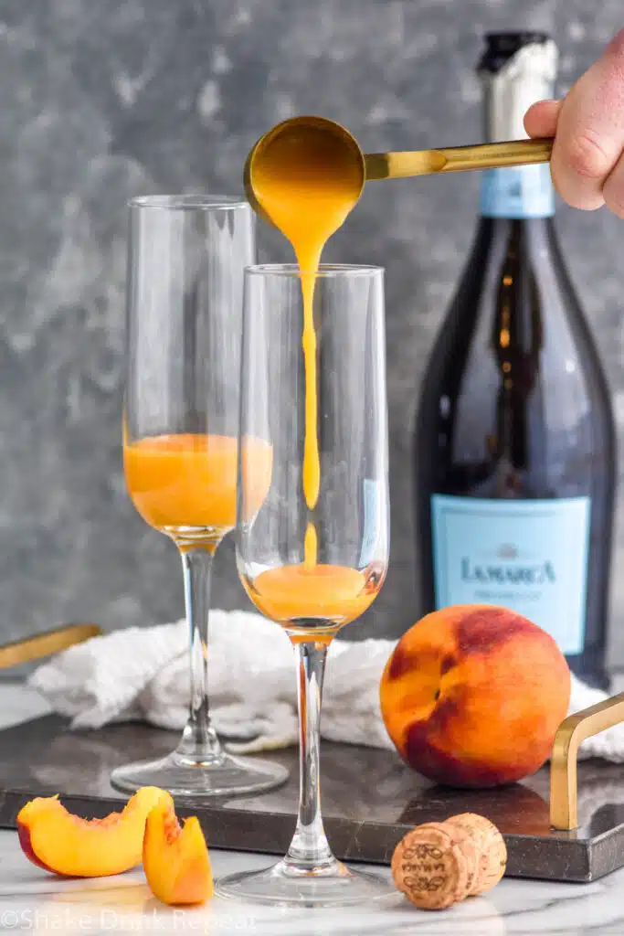man's hand pouring peach puree into a champagne flute to make a peach Bellini. Fresh peach and bottle of champagne sitting in background.