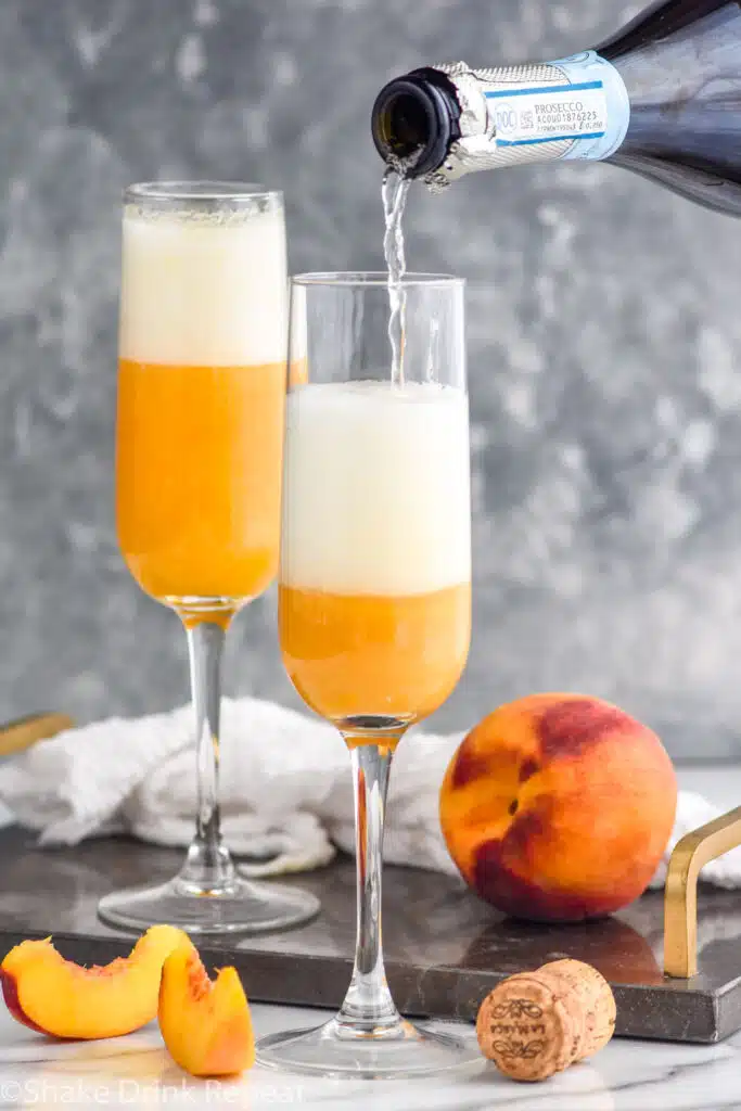 bottle of champagne pouring into a champagne flute to make a Bellini. Fresh peach and peach slices sitting beside.