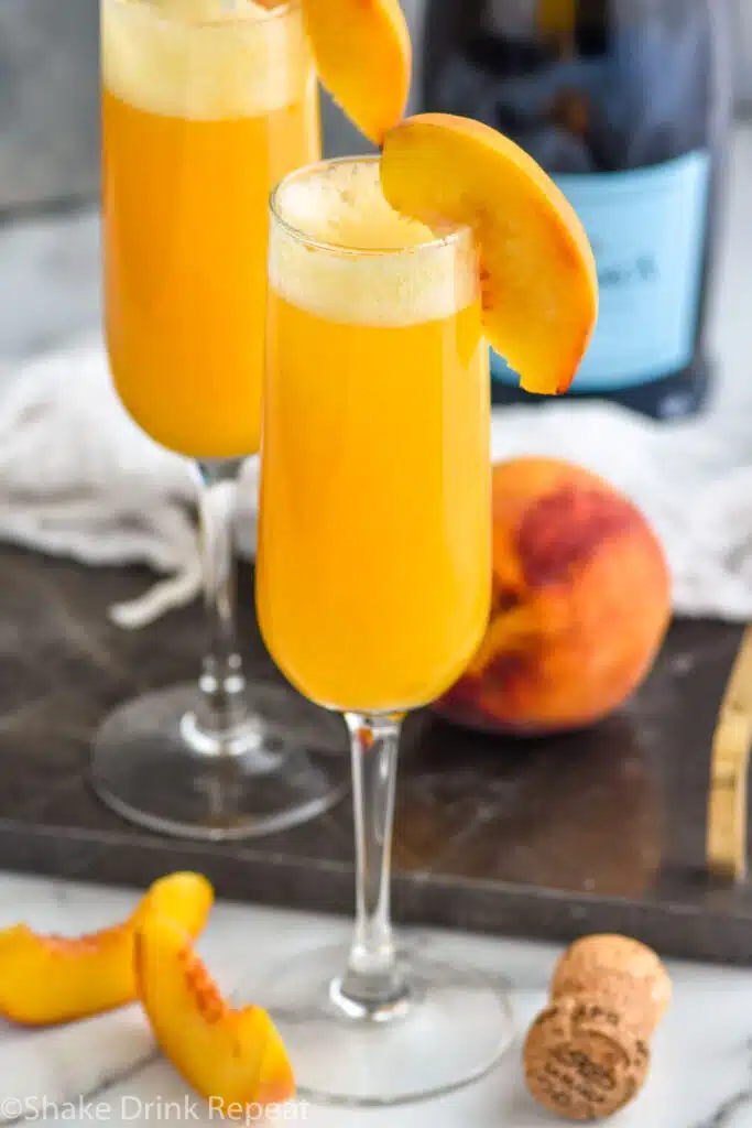 Two glasses of Bellini with fresh peach, peach slices, and bottle of champagne surrounding.