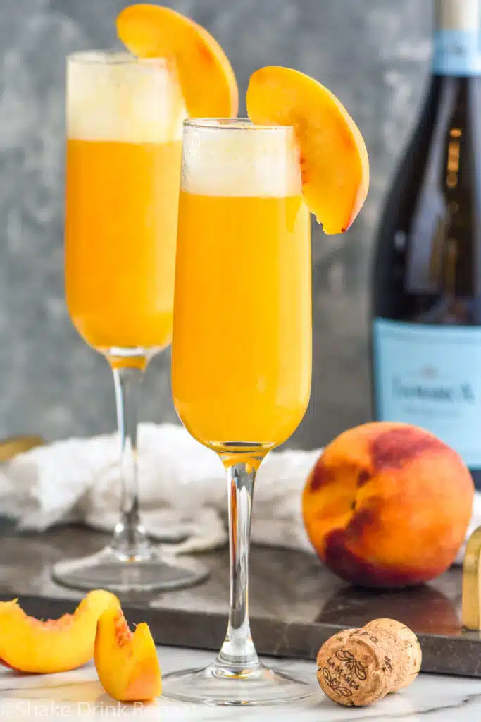 Two glasses of Bellini cocktails garnished with a slice of fresh peach. Bottle of champagne and peach sitting in background.