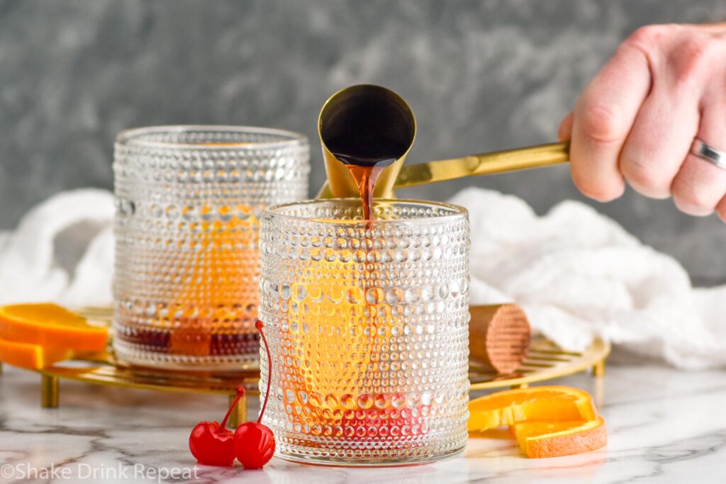 Side view of person's hand pouring cocktail jigger of ingredients into tumbler for Cherry Old Fashioned recipe.