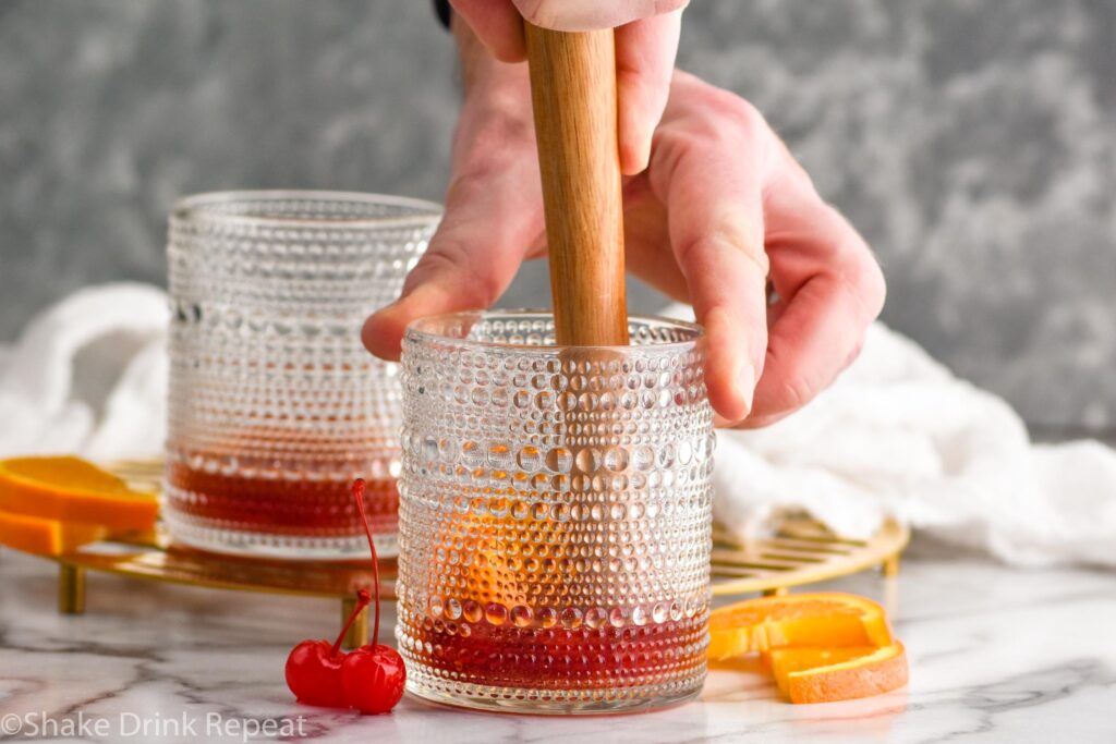 Side view of person muddling ingredients in tumbler for Cherry Old Fashioned recipe. Orange slices and cherries beside.