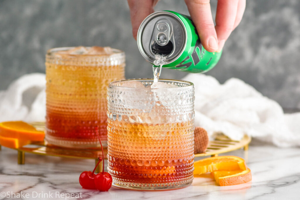 Side view of person's hand pouring lemon lime soda into tumbler for Cherry Old Fashioned recipe. Orange slices and cherries beside.