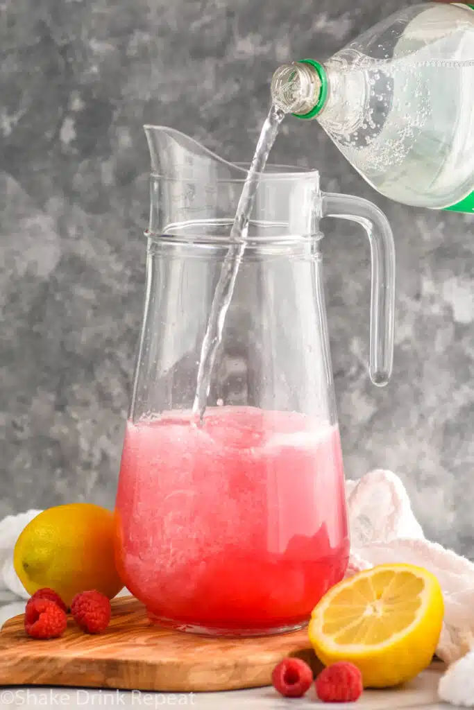 Side view of ingredients being poured into pitcher for Pink Lemonade Vodka Punch recipe. Lemons and raspberries beside.