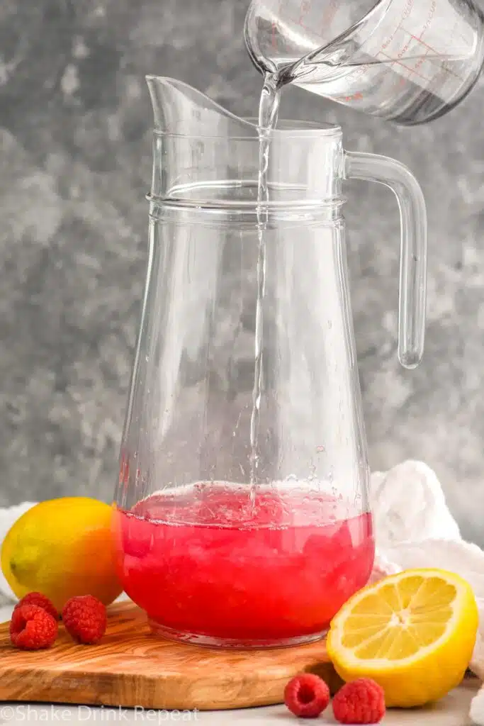 Side view of ingredients being poured into pitcher for Pink Lemonade Vodka Punch recipe. Lemons and raspberries beside.