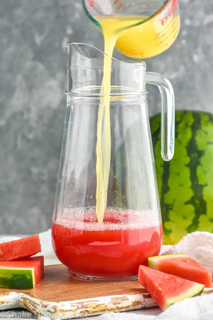 Measuring cup of ingredient being poured into pitcher for Watermelon rum punch recipe. Watermelon beside