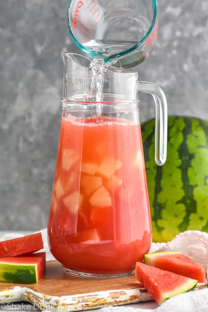 Measuring cup of ingredients being poured into pitcher for Watermelon rum punch recipe. Watermelon beside.