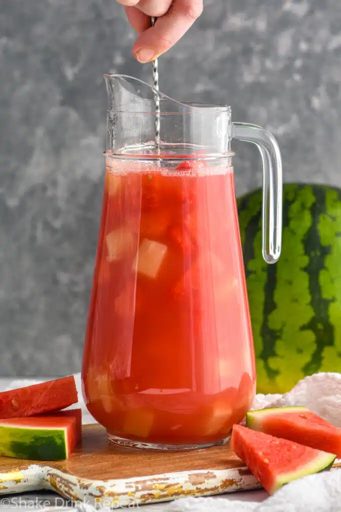 Person's hand stirring pitcher of Watermelon rum punch. Watermelon beside