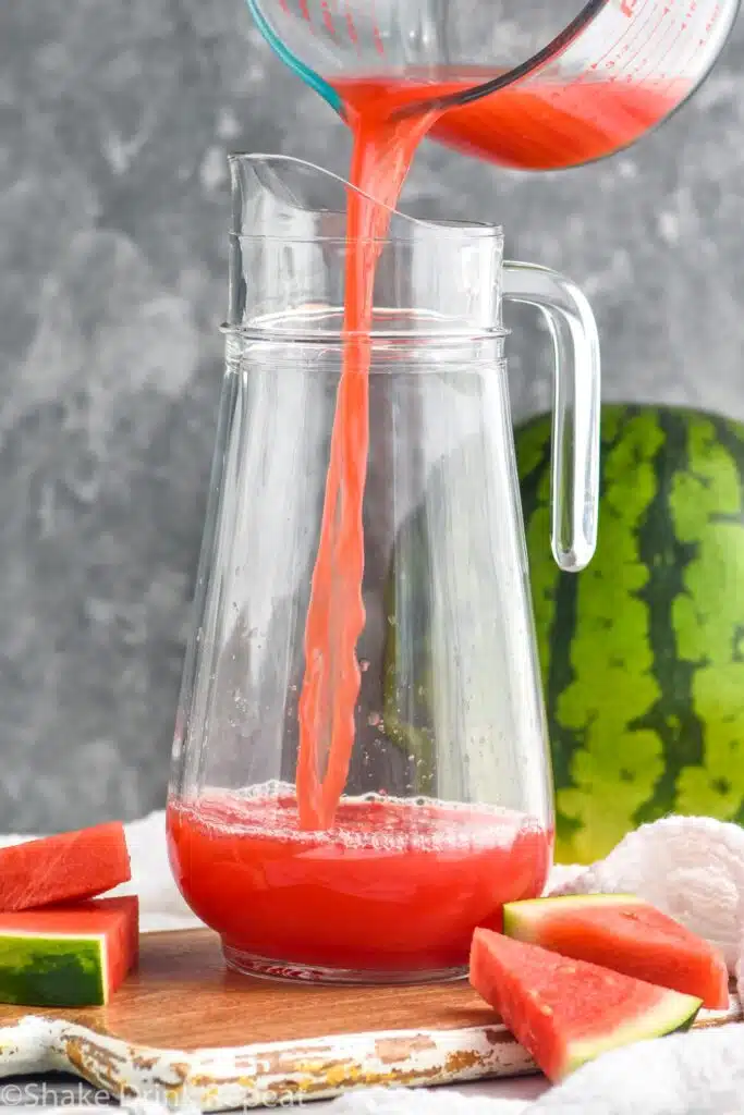 Measuring cup of ingredient being poured into pitcher for Watermelon rum punch recipe. Watermelon beside