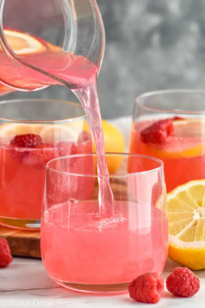 Pitcher of Pink Lemonade Vodka Punch being poured into glasses. Lemon and raspberries beside for garnish
