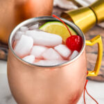 Cherry Moscow Mule served in copper mugs with ice, lime wedge, and cherry as garnish. Cocktail jigger and more cherries beside.