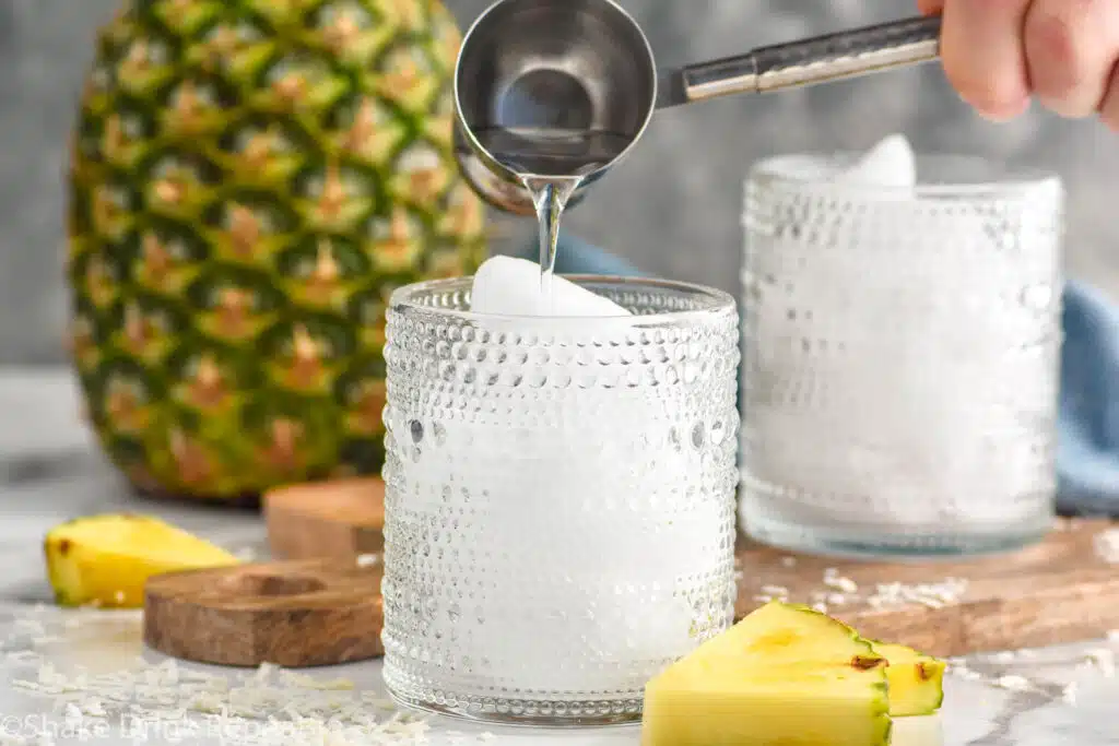 Man's hand pouring cocktail jigger of vodka into a glass to make a Pina Colada Vodka Soda. Fresh pineapple and glass of ice sitting in background