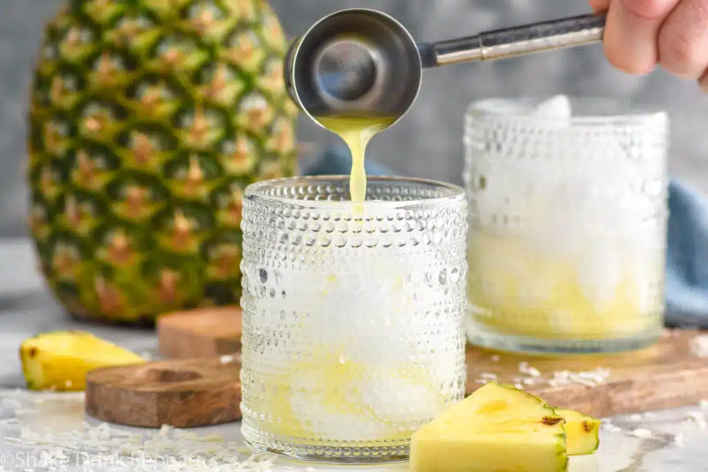 Man's hand pouring cocktail jigger of pineapple juice into a glass to make a Pina Colada Vodka Soda. Fresh pineapple and glass of ice and pina colada vodka soda ingredients sitting in background