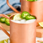 Jalapeno Moscow Mules served in copper mugs with ice, sliced jalapenos, and lime wedge as garnish. More sliced jalapenos and lime wedges beside copper mugs.