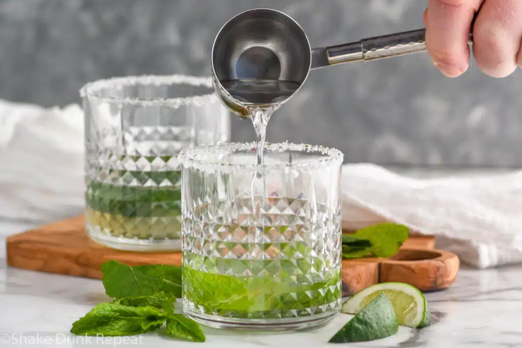 man's hand pouring cocktail jigger of tequila into a glass of Mojito Margarita ingredients to make a Mojito Margarita. Glass with mint leaves, fresh mint leaves, and lime wedges surrounding.
