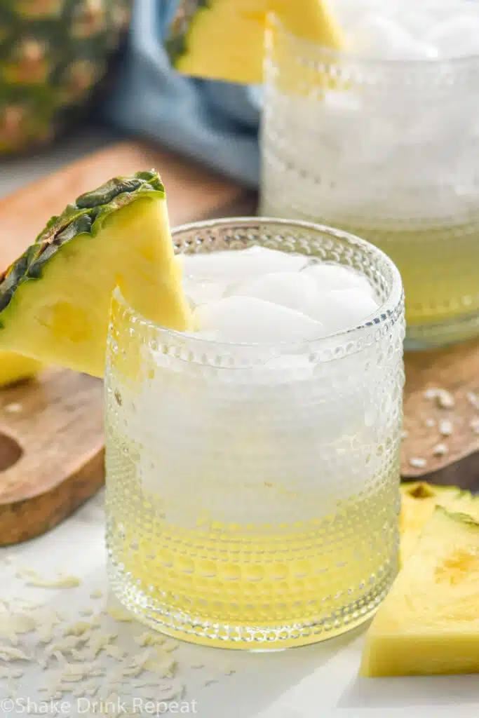 glass of Pina Colada Vodka Soda with ice garnished with a slice of pineapple. Fresh pineapple and glass of Pina Colada Vodka Soda sitting behind