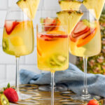 Four wine glasses of Tropical Margarita Sangria garnished with a slice of pineapple. Fresh strawberries and kiwi sitting beside.