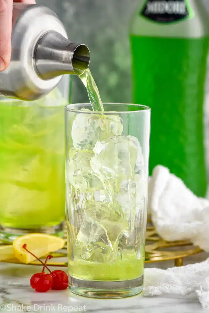 cocktail shaker of tokyo tea ingredients pouring into a glass of ice. Bottle of midori, glass of Tokyo Tea, lemon slices, and cherries sitting in background