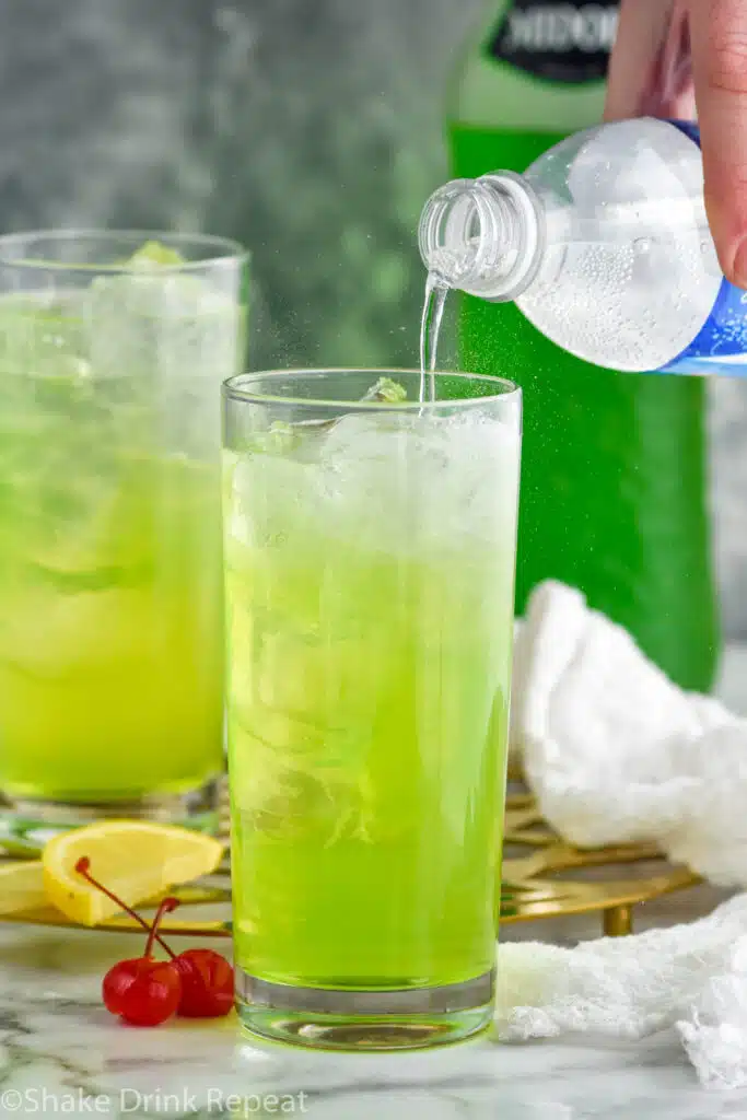 Man's hand pouring bottle of club soda into a glass of Tokyo Tea ingredients and ice. Glass of Tokyo Tea, bottle of Midori, lemon slices, and cherries sitting behind