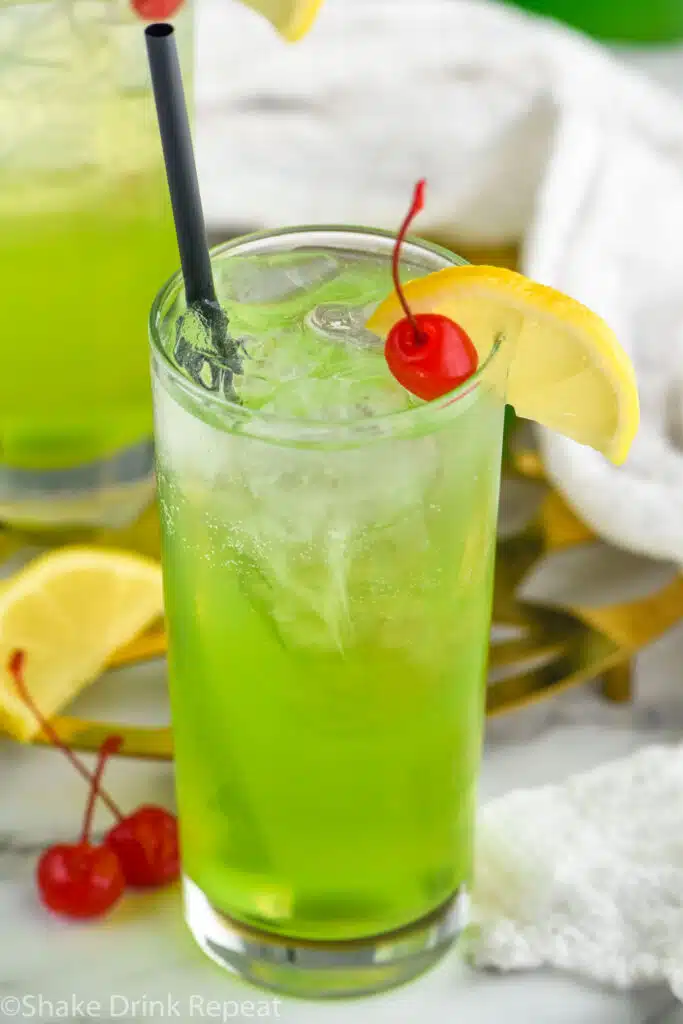 Glass of Tokyo Tea with ice, straw, lemon slice, and cherry. Lemon slice and cherries sitting beside
