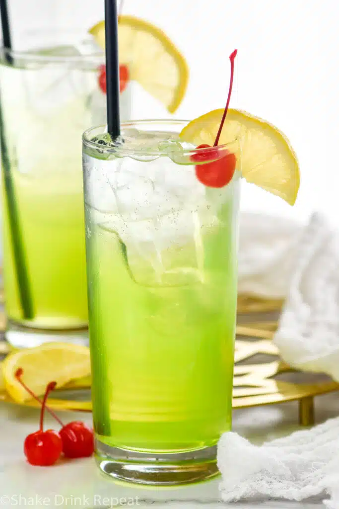 Two glasses of Tokyo Tea with ice, straw, lemon slices, and cherry. Two cherries and lemon slice sitting beside.